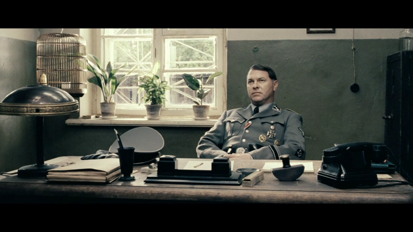 "I will arrange another kind of death for you." The ultimate degradation of humanity. (And two other queries: Is it a coincident that those three plants are conspicuously placed behind the German office and what is the symbolism of the bird?